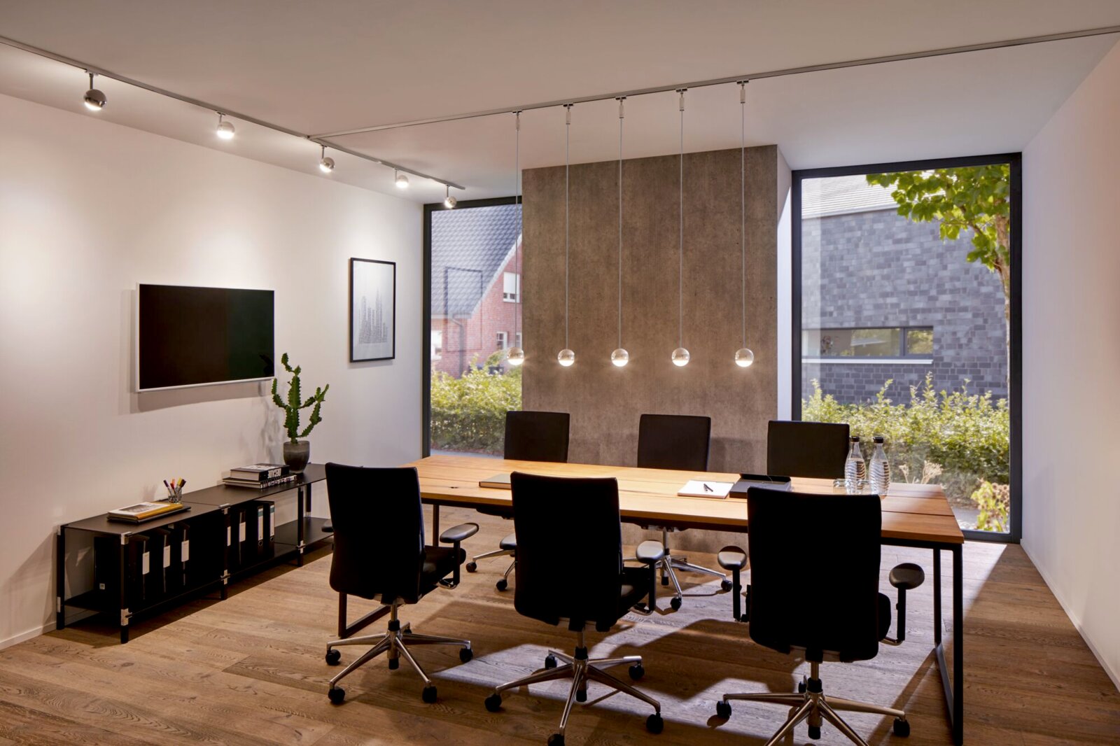 Moderne kantoorverlichting met kabelophanging, zijaanzicht op een vergadertafel met vijf stoelen en grote ramen op de achtergrond.