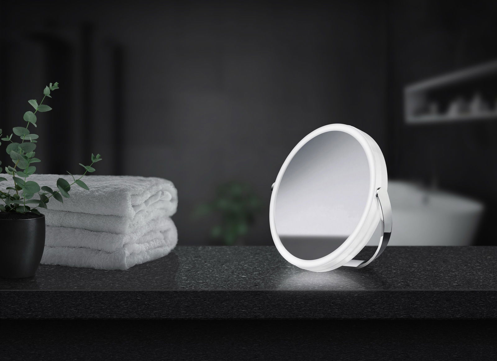 Verchromter Kosmetikspiegel mit LED-Beleuchtung auf einer dunklen Badezimmerablage, seitliche Perspektive