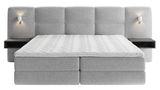 Frontale Ansicht eines grauen Boxspringbetts mit gepolstertem Kopfteil, integrierten Nachttischen und Wandlampen.