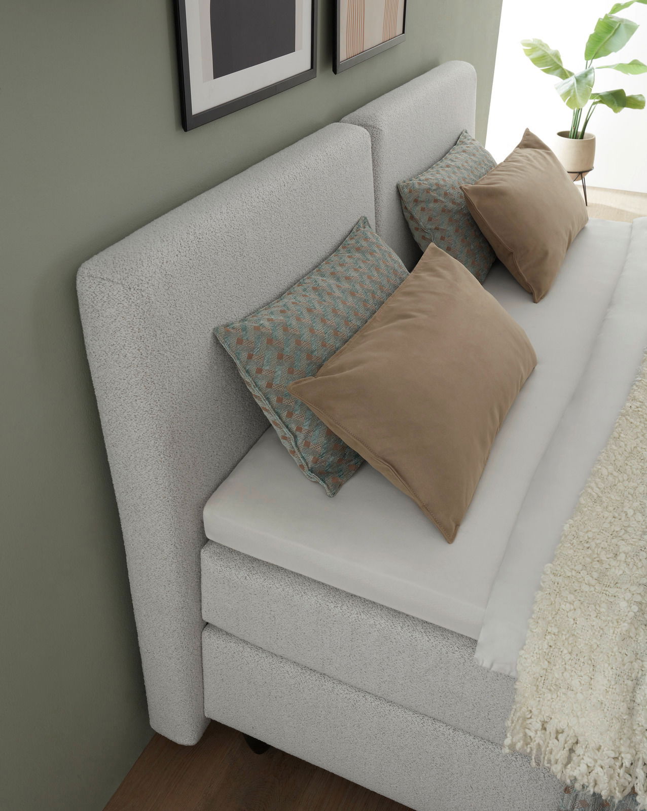 Boxspringbett in seitlicher Perspektive mit grauem Kopfteil, dekoriert mit Kissen in Beige und gemustertem Design, vor einer grünen Wand mit Pflanzen und Bildern.