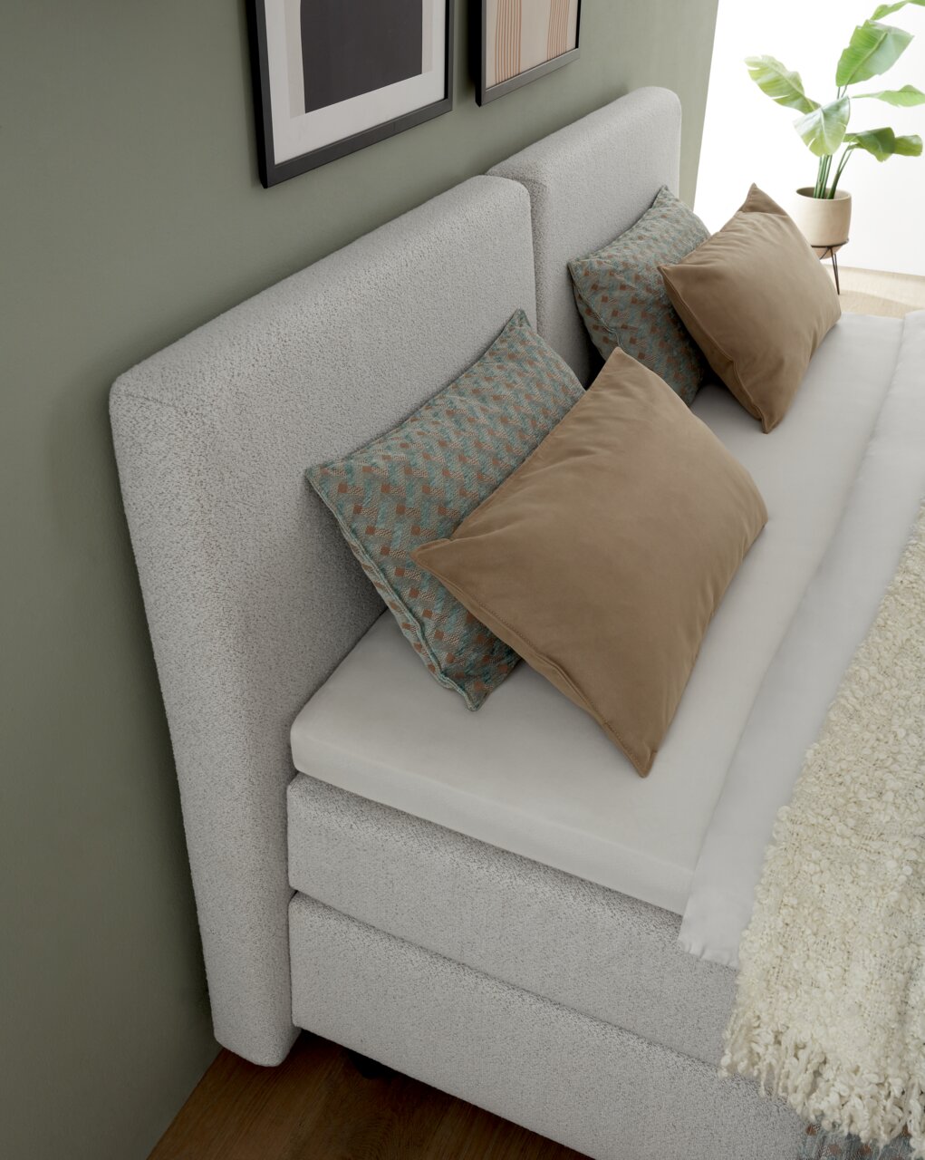 Boxspringbett in seitlicher Perspektive mit grauem Kopfteil, dekoriert mit Kissen in Beige und gemustertem Design, vor einer grünen Wand mit Pflanzen und Bildern.