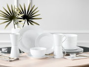 Weißes, minimalistisches Porzellan-Geschirrset mit Tellern, Schalen und Tassen auf einem Tisch arrangiert, perspektivisch in frontaler Ansicht mit leichter Schrägansicht.