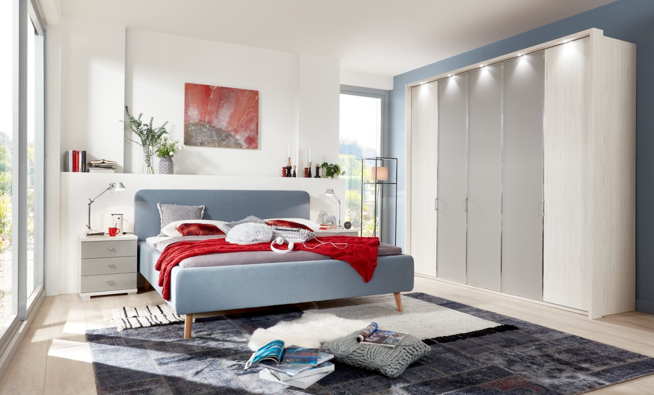 Modernes Polsterbett in einem hellen Schlafzimmer, seitliche Perspektive, mit blauer Bettumrandung und roter Bettdecke, neben einem Nachttisch und einem großen Kleiderschrank.