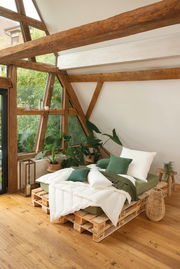 Gemütliches Schlafzimmer mit einem Solobett aus Paletten, dekoriert mit grünen Kissen und Decken, umgeben von Pflanzen, in einem Raum mit großen Fenstern und Holzbalken, aus der Seitenperspektive aufgenommen.