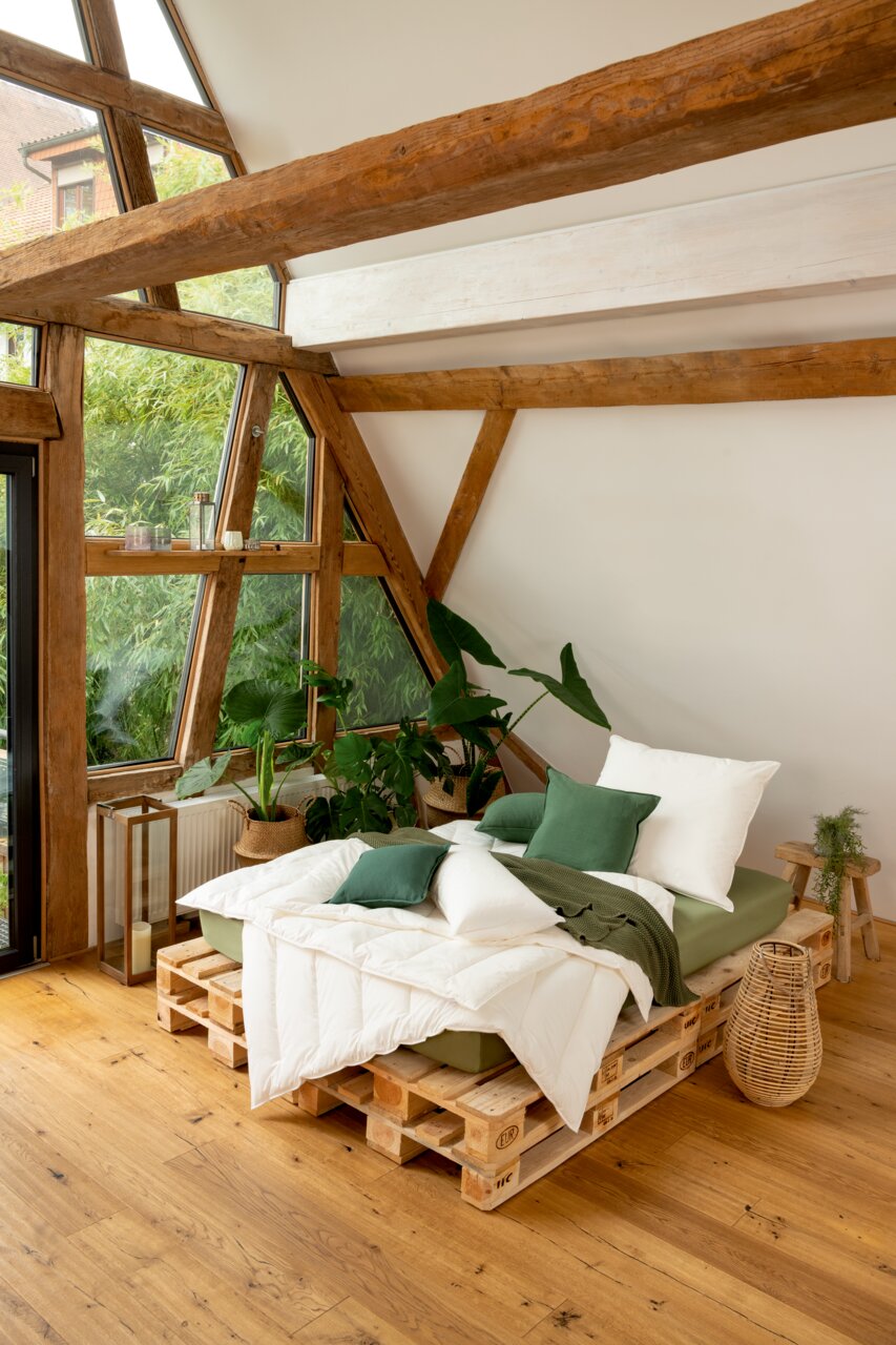 Gemütliches Schlafzimmer mit einem Solobett aus Paletten, dekoriert mit grünen Kissen und Decken, umgeben von Pflanzen, in einem Raum mit großen Fenstern und Holzbalken, aus der Seitenperspektive aufgenommen.