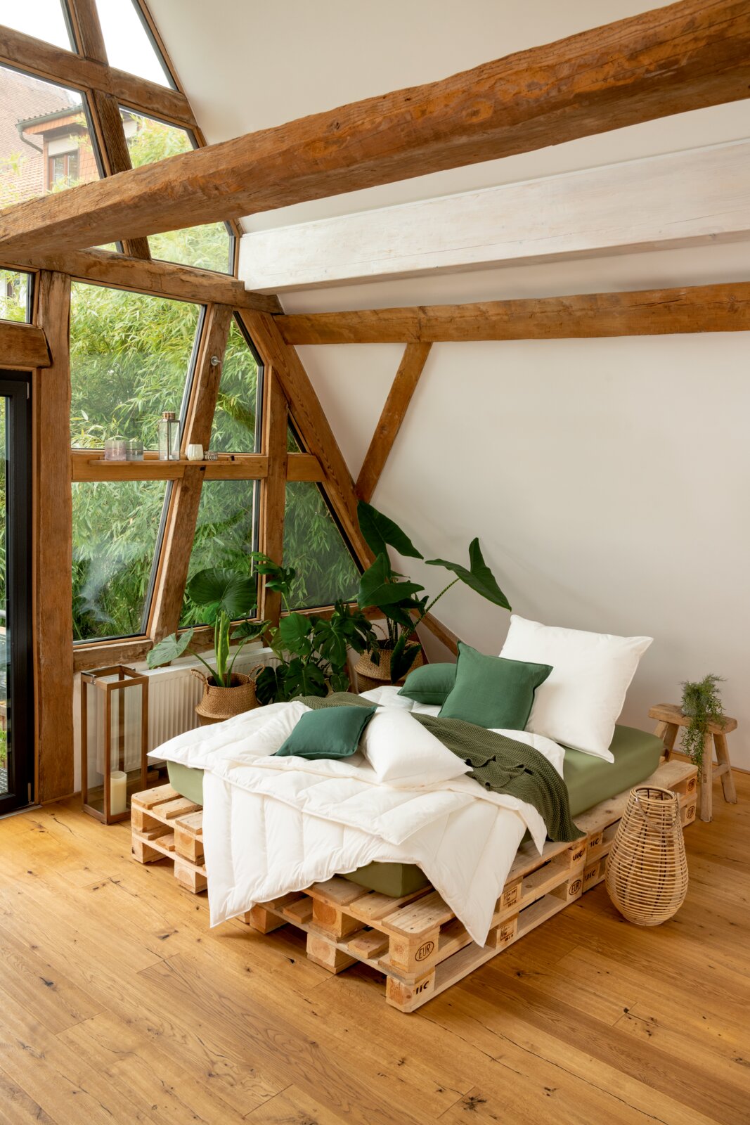 Gemütliches Schlafzimmer mit einem Solobett aus Paletten, dekoriert mit grünen Kissen und Decken, umgeben von Pflanzen, in einem Raum mit großen Fenstern und Holzbalken, aus der Seitenperspektive aufgenommen.