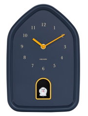 Moderne Wanduhr in Hausform mit gelben Zeigern und einer kleinen Figur in der unteren Mitte, Frontalansicht.