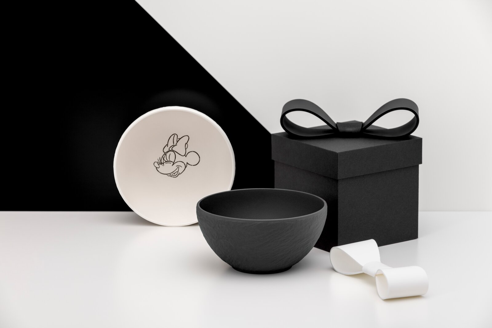 Schwarz-weißes Schalen-Set mit Minnie-Maus-Motiv, daneben schwarze Geschenkbox mit Schleife und weißes Band, aufgenommen in leichter Seitenansicht (3/4-Perspektive) vor minimalistischem Hintergrund.