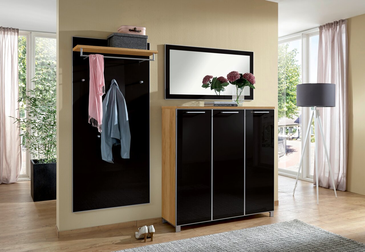 Modernes Garderoben-Set in einem hellen Raum mit großem Fenster, bestehend aus einem schwarzen Schrank, einem Spiegel und einer Garderobenleiste mit Ablage. Perspektive von vorne.