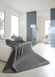 Graue Wohndecke 150x200 cm auf einem Sofa in einem modernen, hellen Wohnzimmer mit großen Fenstern und minimalistischer Dekoration, Perspektive von vorne.