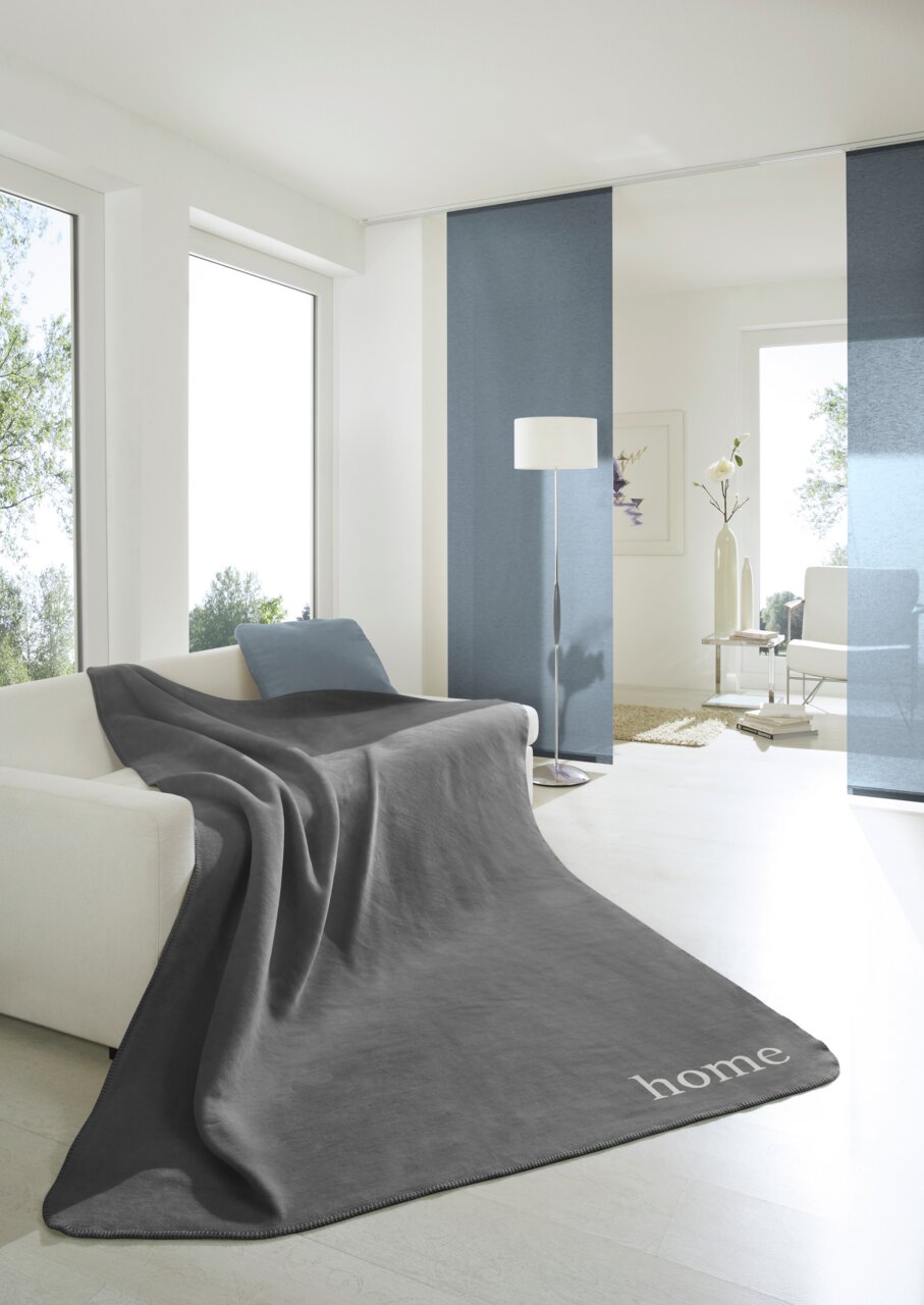 Graue Wohndecke 150x200 cm auf einem Sofa in einem modernen, hellen Wohnzimmer mit großen Fenstern und minimalistischer Dekoration, Perspektive von vorne.