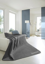 Graue Wohndecke 150x200 cm auf einem Sofa in einem modernen, hellen Wohnzimmer mit großen Fenstern und minimalistischer Dekoration, Perspektive von vorne.