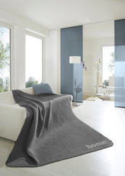 Graue Wohndecke 150x200 cm auf einem Sofa in einem modernen, hellen Wohnzimmer mit großen Fenstern und minimalistischer Dekoration, Perspektive von vorne.