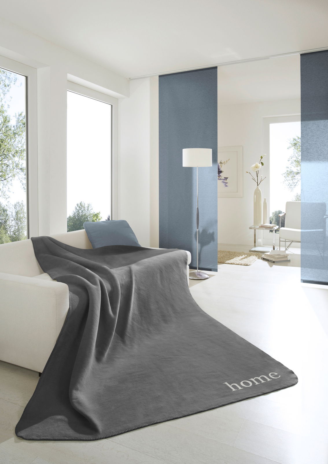 Graue Wohndecke 150x200 cm auf einem Sofa in einem modernen, hellen Wohnzimmer mit großen Fenstern und minimalistischer Dekoration, Perspektive von vorne.