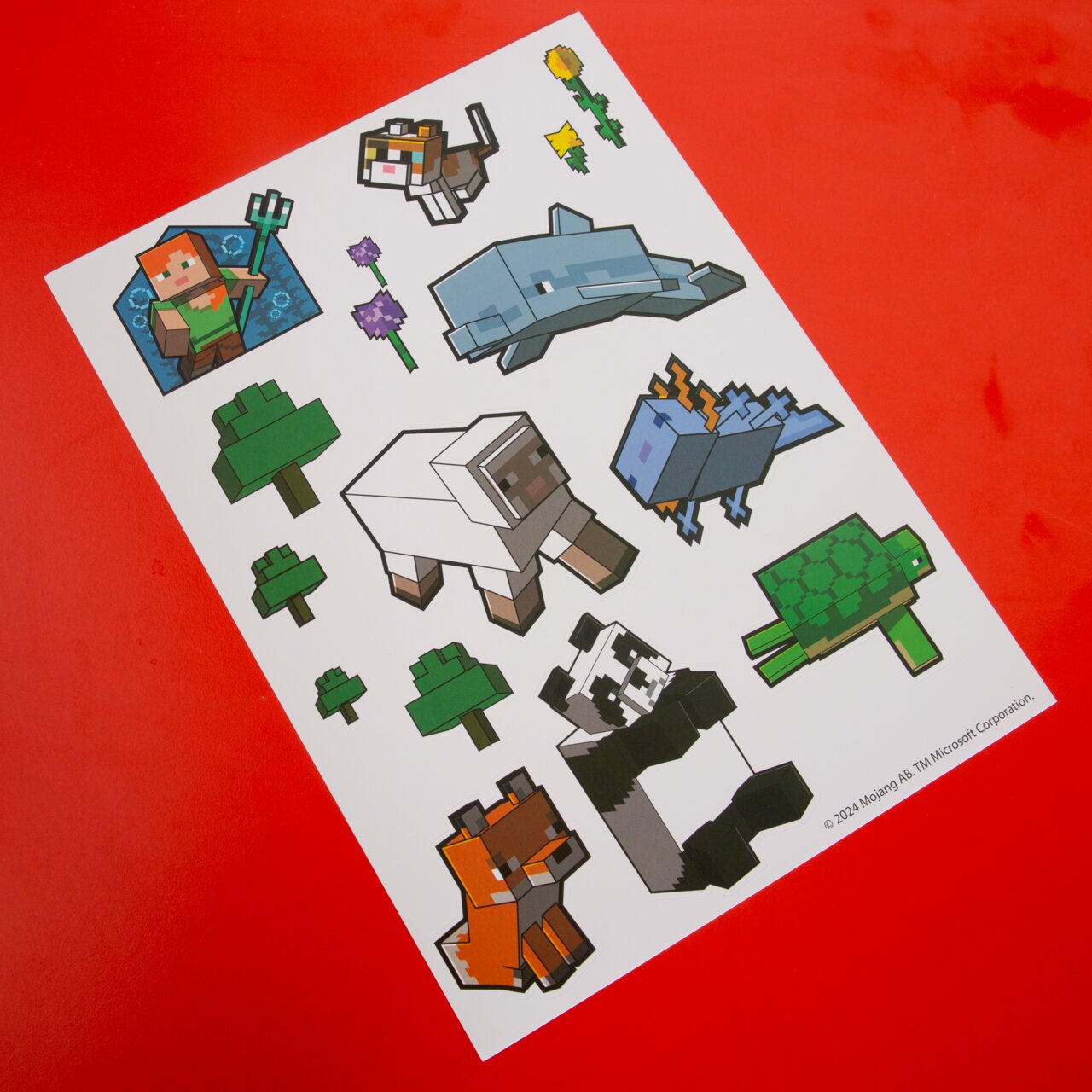 Schräg von oben fotografierter Stickerbogen mit Minecraft-Motiven (u. a. Delfin, Panda, Schildkröte, Schaf, Katze, Fuchs, Bäume und Blumen) auf rotem Hintergrund.