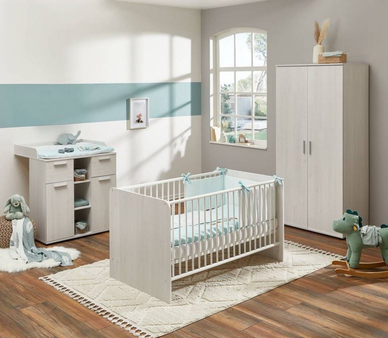 Kinderzimmer mit hellem Holzunterstellregal, Wickelkommode und Gitterbett, aus der Perspektive eines Fensters mit Tageslicht.