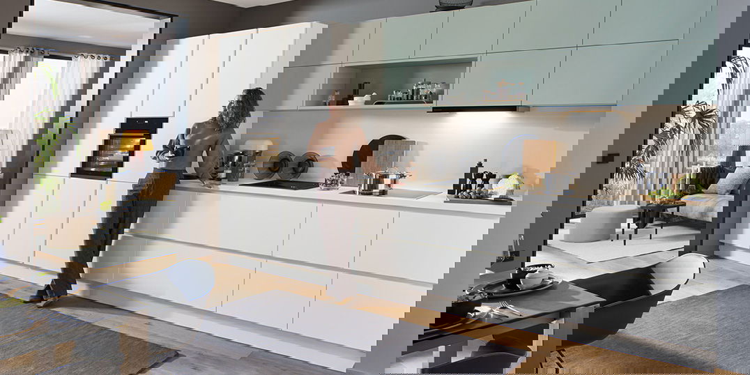 Eine moderne Küche mit mintgrünen Oberschränken und einem großen Fenster. Eine Frau lehnt an einem Hochschrank.