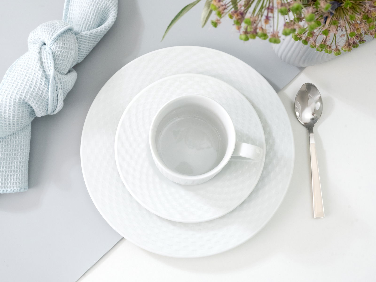 Kaffeeservice bestehend aus Tasse, Untertasse und Teller in eleganter weißer Optik, von oben fotografiert, mit Besteck und Serviette arrangiert.