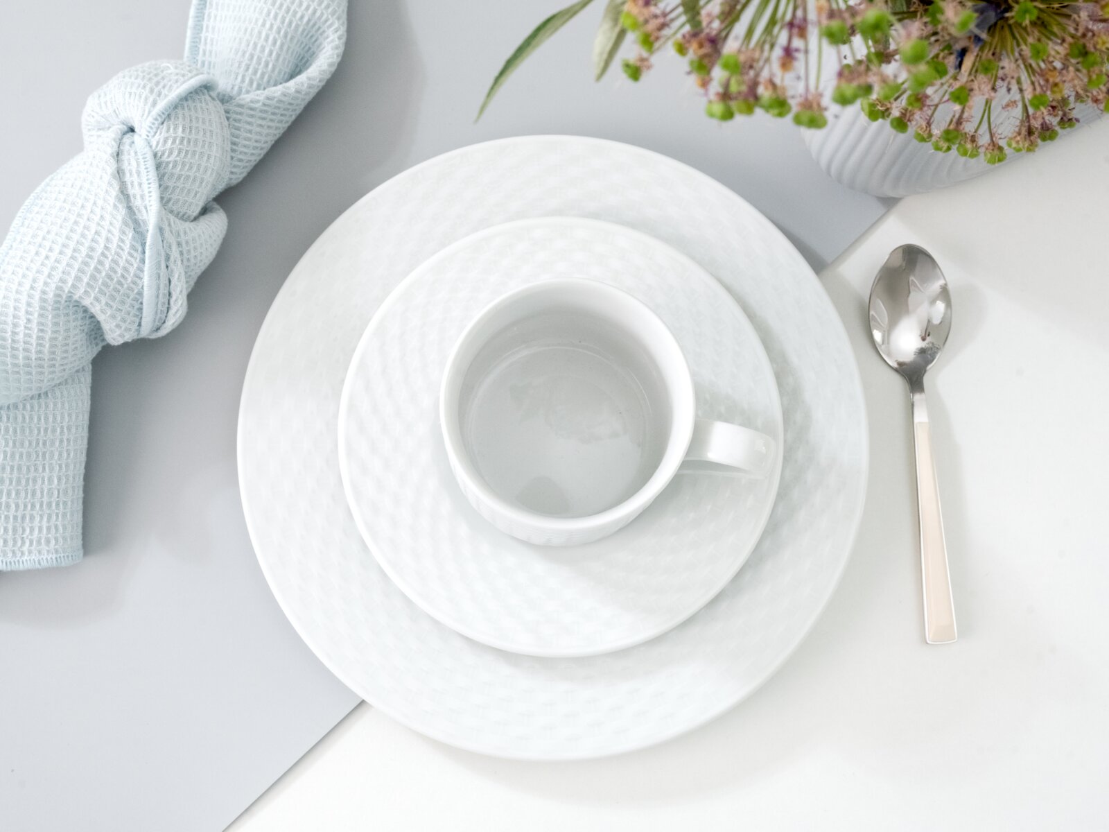 Kaffeeservice bestehend aus Tasse, Untertasse und Teller in eleganter weißer Optik, von oben fotografiert, mit Besteck und Serviette arrangiert.