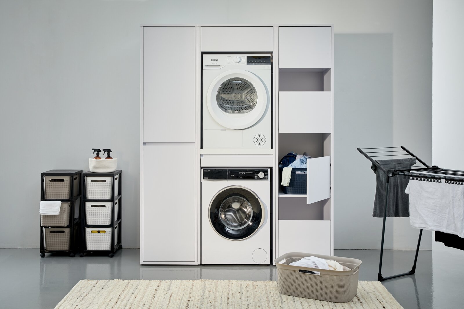 Waschmaschinenschrank mit integriertem Trockner und Waschmaschine, umgeben von Stauraum und Wäschezubehör, frontal betrachtet.