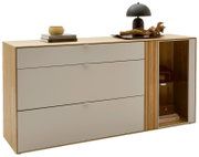 Modernes Sideboard aus hellem Holz mit drei breiten, graubeigen Schubladen und rechter Vitrine mit Glastür, dekoriert mit Büchern und Lampe, in frontaler leicht schräger Perspektive von links auf weißem Hintergrund.