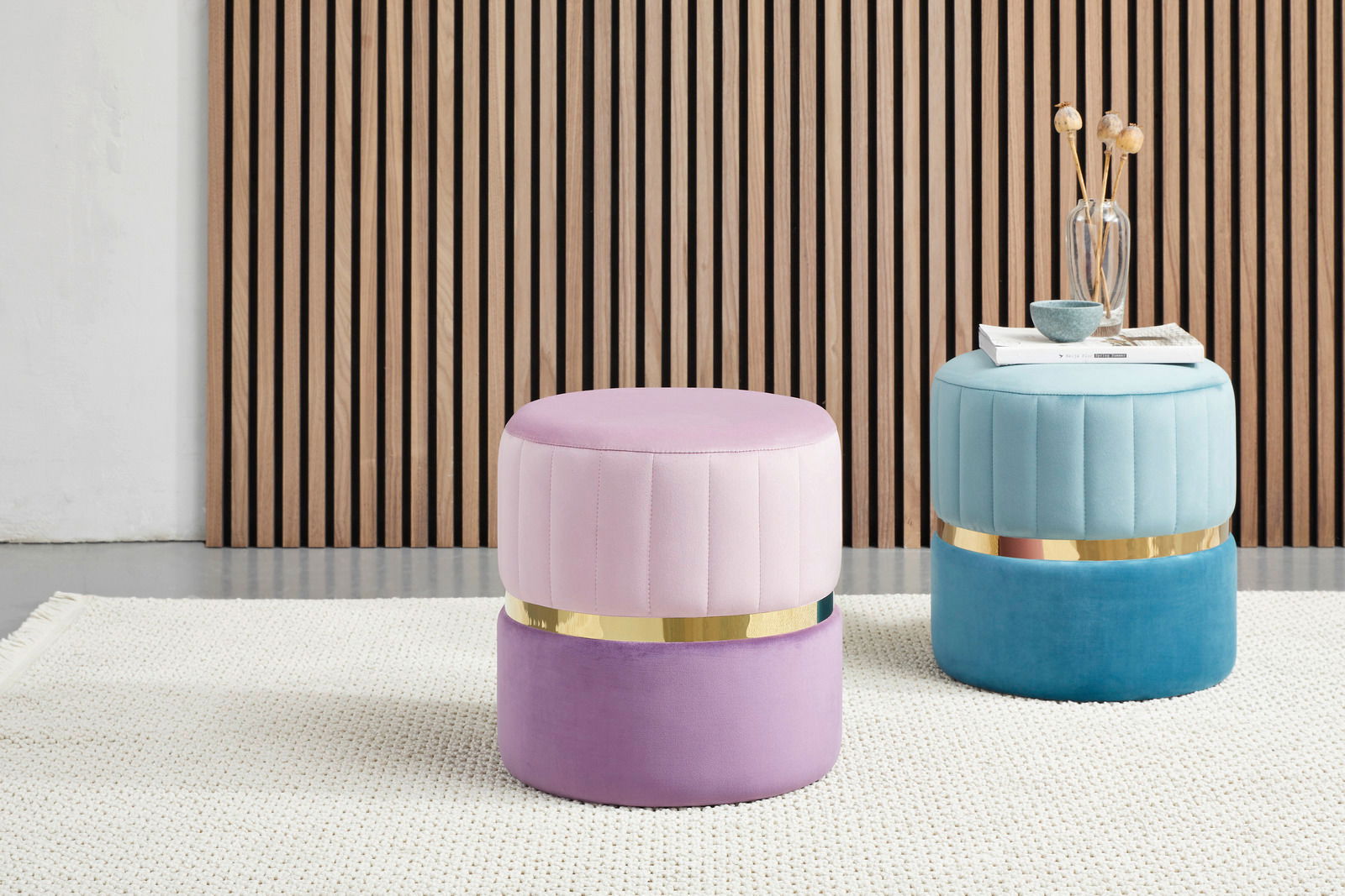 Zwei moderne Hocker in Pastellfarben, einer in Rosa und Lila, der andere in Türkis und Blau, mit goldenen Akzenten, auf einem hellen Teppich vor einer Holzlamellenwand. Perspektive: Frontalansicht.