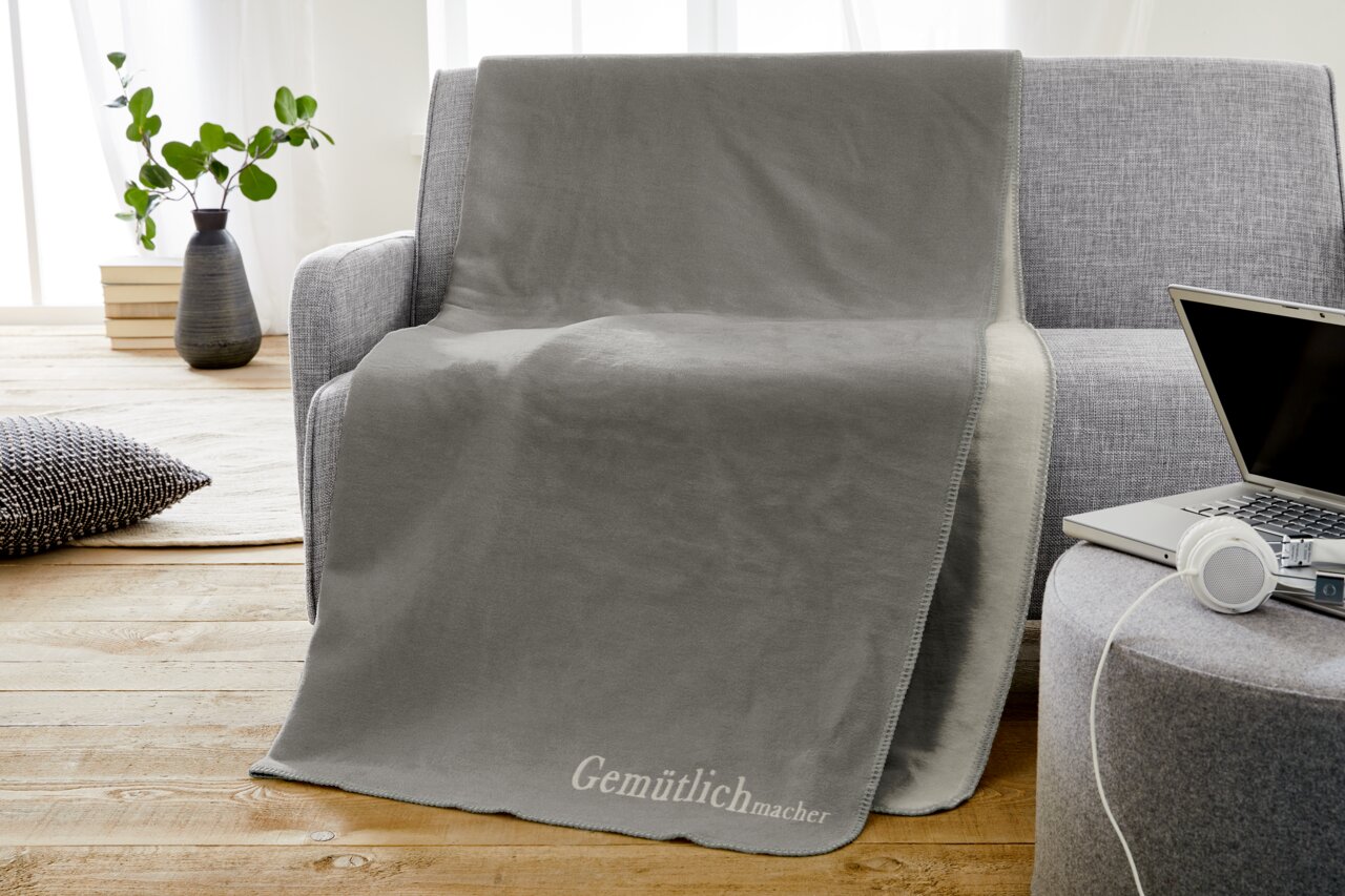 Graue Jacquard-Decke 150x200cm auf einem grauen Sofa drapiert, mit der Aufschrift 'Gemütlichmacher' in einem modernen Wohnzimmer mit Holzboden und Pflanzen im Hintergrund, seitliche Perspektive.