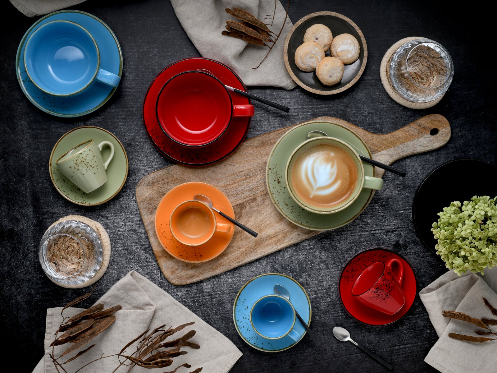 Buntes Keramik-Tassen- und Untertassen-Set in Rot, Blau, Grün und Orange mit Löffeln, auf dunklem Tisch arrangiert; Draufsicht/Flatlay, mit Holzbrett, Cappuccino-Latte-Art und kleinen Keksen.