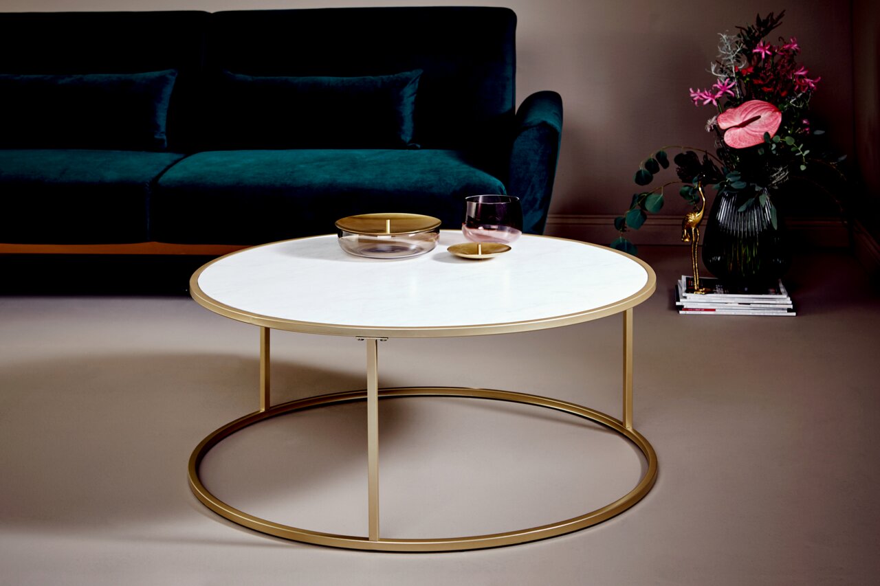 Runder Couchtisch mit weißer Tischplatte und goldfarbenem Gestell, seitliche Perspektive, im Wohnzimmer mit grünem Sofa und Blumenarrangement.