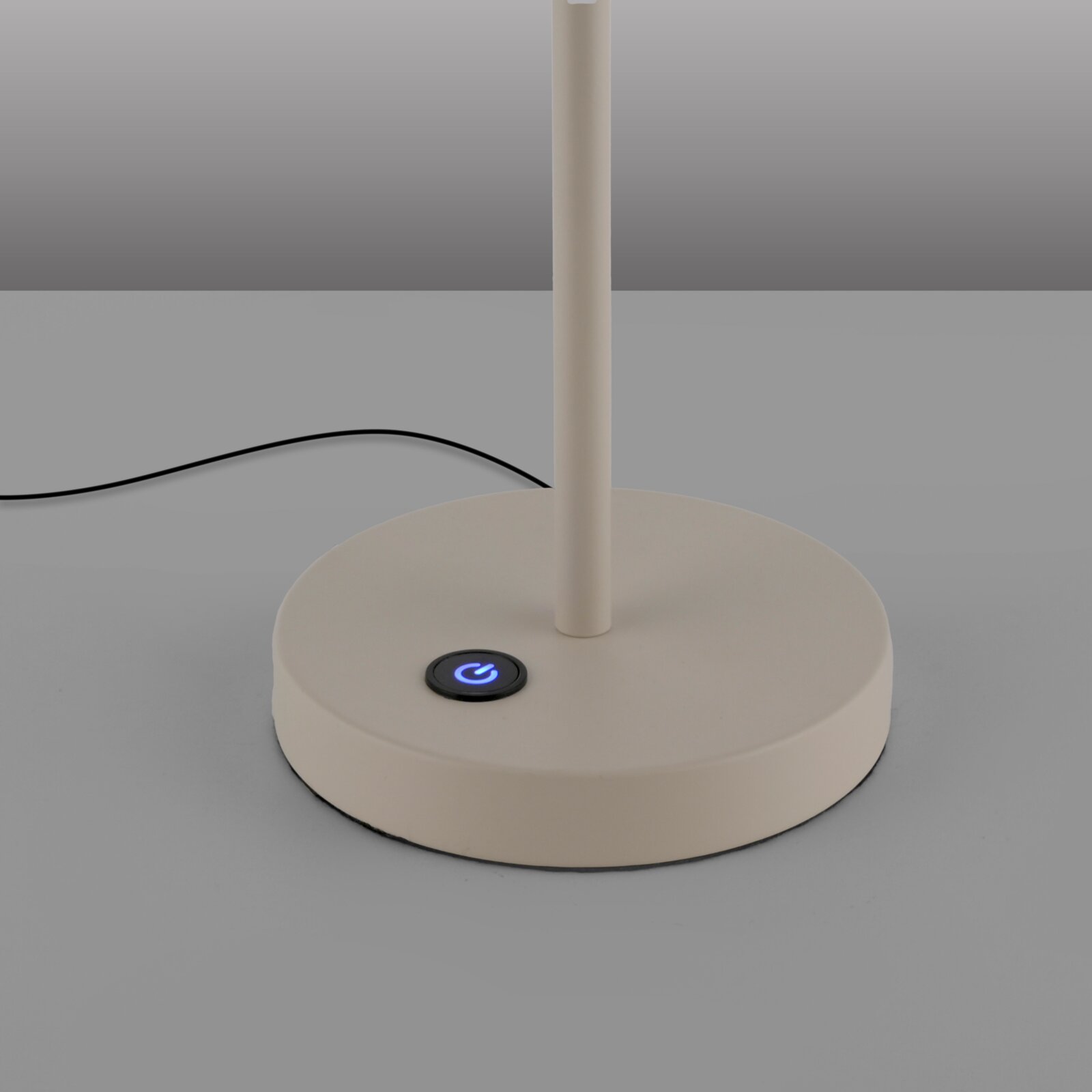 Nahaufnahme des runden Standfußes einer weißen Tischlampe mit blau beleuchtetem Touch-Ein-/Aus-Schalter und schwarzem Kabel, fotografiert frontal aus leicht erhöhter Perspektive