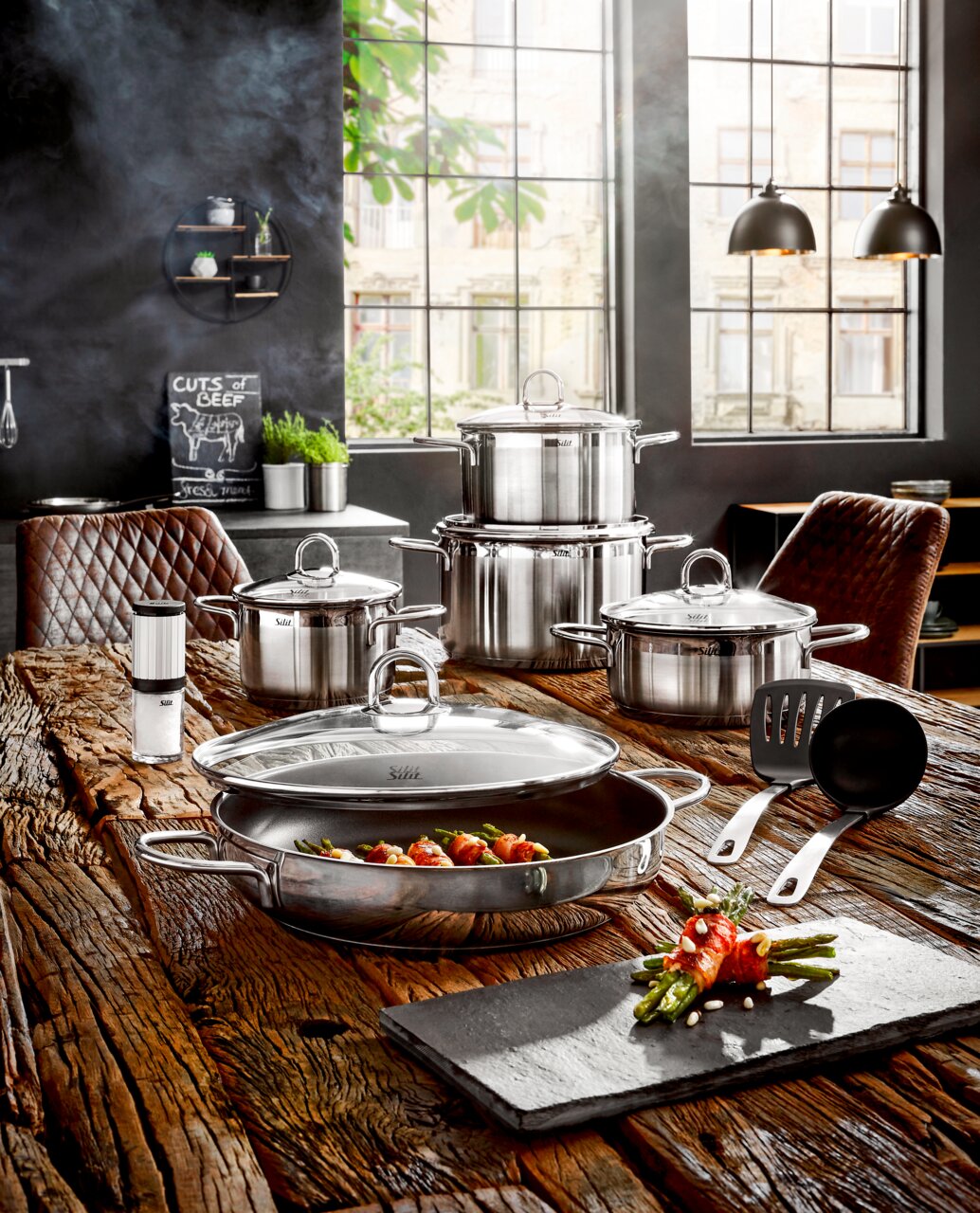 Elegantes 9-teiliges Topf-Set Diamant aus Edelstahl, präsentiert auf einem rustikalen Holztisch in einer modernen Küche mit großen Fenstern im Hintergrund. Perspektive von vorne.