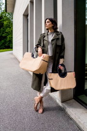 Frau lehnt an einer Wand und hält zwei große, beige Taschen mit schwarzen Griffen. Die Taschen sind aus Raffia-Material und haben ein modernes Design. Perspektive von vorne.