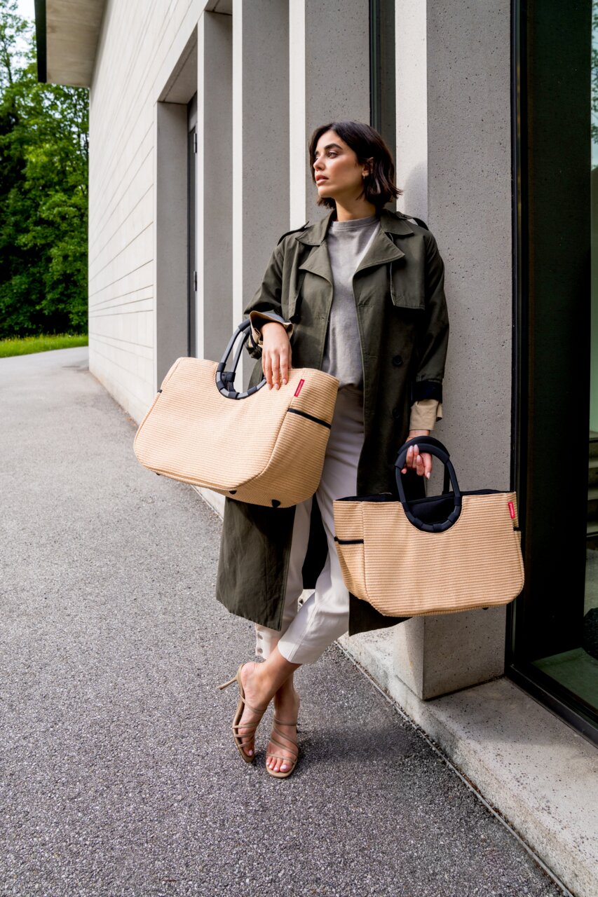 Frau lehnt an einer Wand und hält zwei große, beige Taschen mit schwarzen Griffen. Die Taschen sind aus Raffia-Material und haben ein modernes Design. Perspektive von vorne.