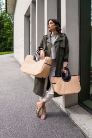 Frau lehnt an einer Wand und hält zwei große, beige Taschen mit schwarzen Griffen. Die Taschen sind aus Raffia-Material und haben ein modernes Design. Perspektive von vorne.