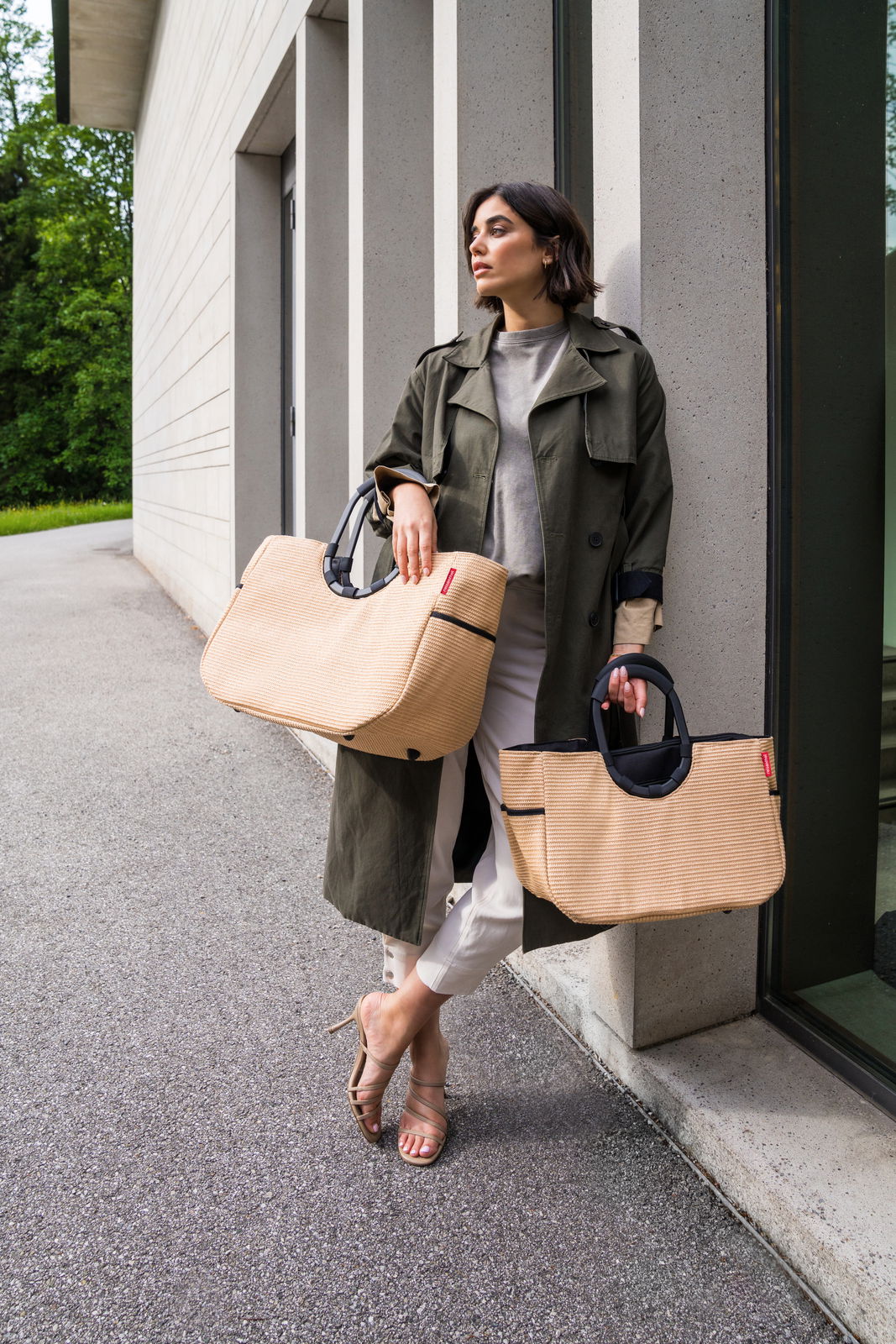 Frau lehnt an einer Wand und hält zwei große, beige Taschen mit schwarzen Griffen. Die Taschen sind aus Raffia-Material und haben ein modernes Design. Perspektive von vorne.