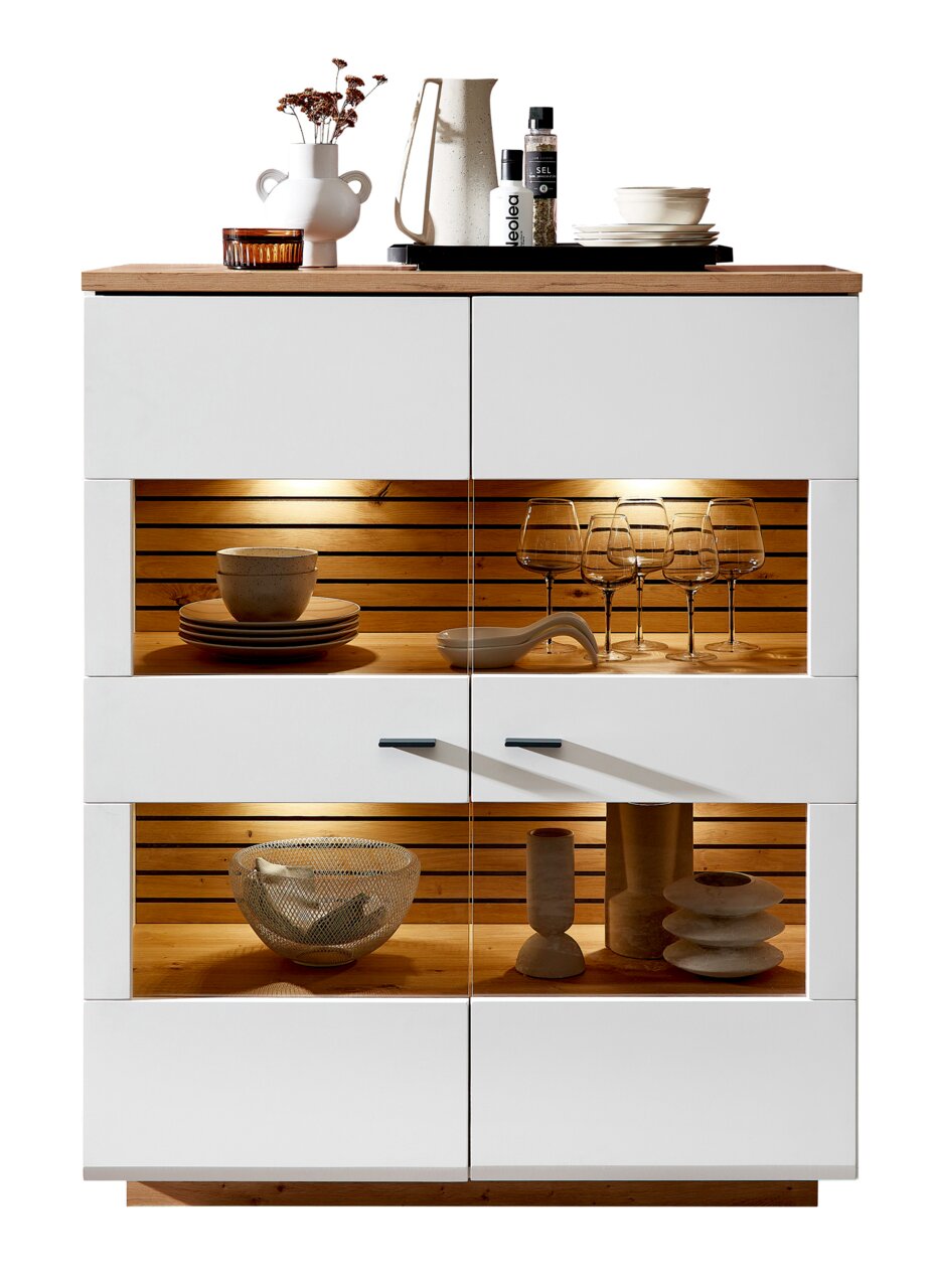 Zurbrüggen Hoog dressoir FRESNO Vooraanzicht van het Zurbrüggen FRESNO-hoogkastje met witte romp, houten details en ingebouwde verlichting, gedecoreerd met servies en glazen.