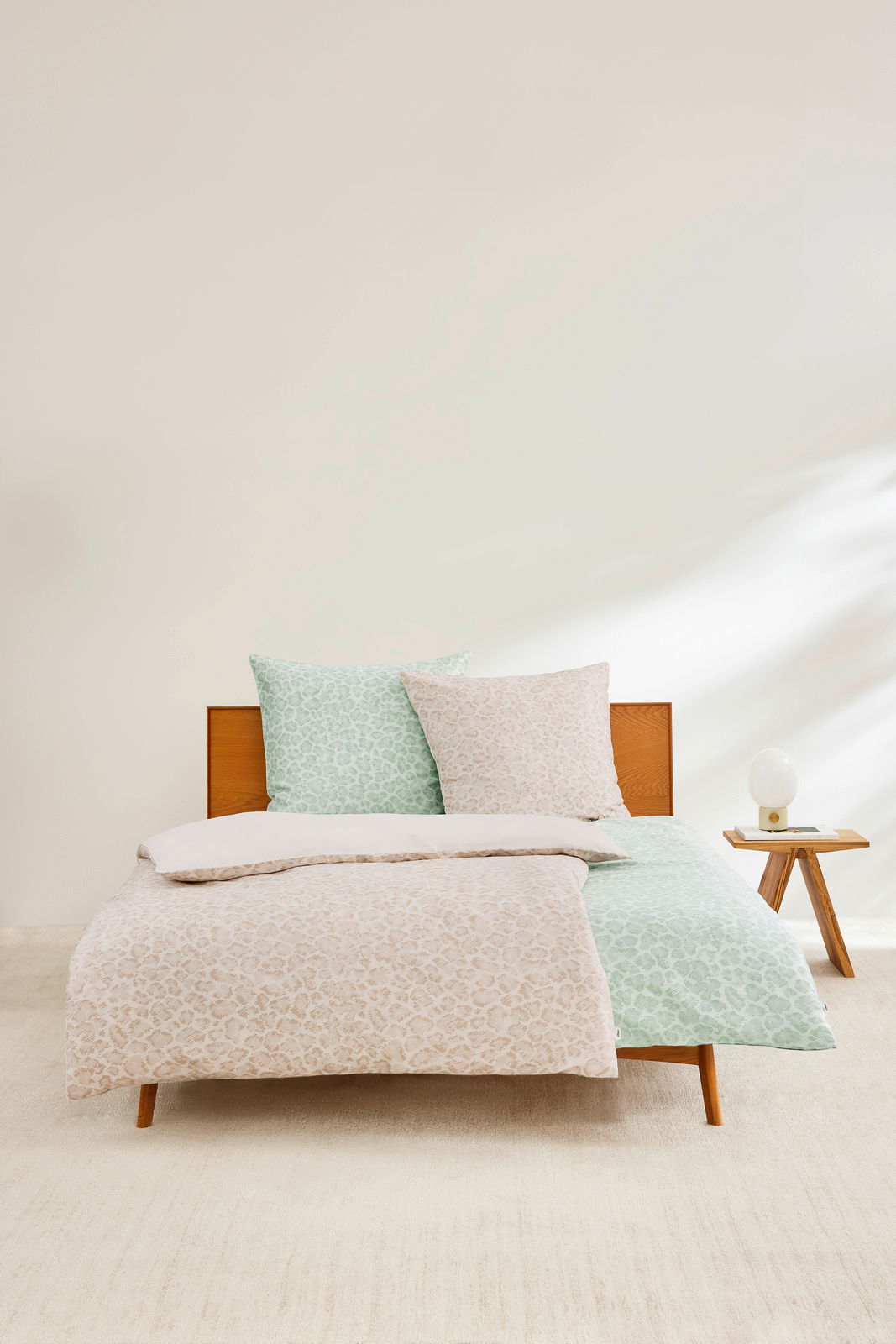 Satinbettwäsche-Set in Beige und Grün mit dezentem Muster, auf einem Holzrahmenbett präsentiert, mit einem Nachttisch und Lampe im Hintergrund, aus der Frontalperspektive fotografiert.