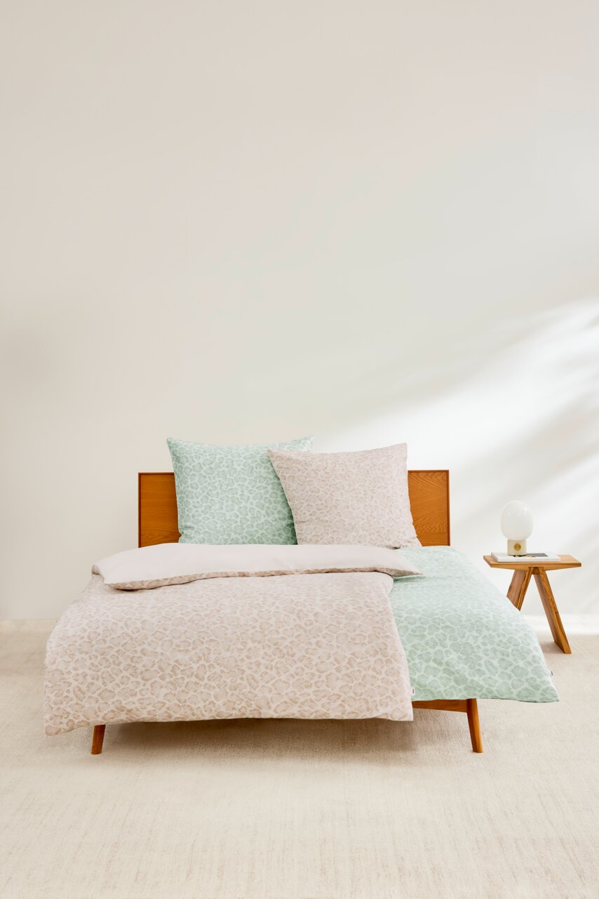 Satinbettwäsche-Set in Beige und Grün mit dezentem Muster, auf einem Holzrahmenbett präsentiert, mit einem Nachttisch und Lampe im Hintergrund, aus der Frontalperspektive fotografiert.