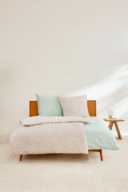 Satinbettwäsche-Set in Beige und Grün mit dezentem Muster, auf einem Holzrahmenbett präsentiert, mit einem Nachttisch und Lampe im Hintergrund, aus der Frontalperspektive fotografiert.