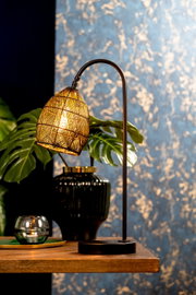 Sfeervolle zwarte tafellamp met gebogen standaard en warm verlichte, gevlochten kap, vanuit schuin vooraanzicht gefotografeerd op een houten tafel.