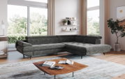 Modernes graues Ecksofa in L-Form mit gepolsterten Armlehnen und schwarzen Metallfüßen in einem hellen Wohnzimmer, seitliche Frontansicht aus leicht schräger Perspektive