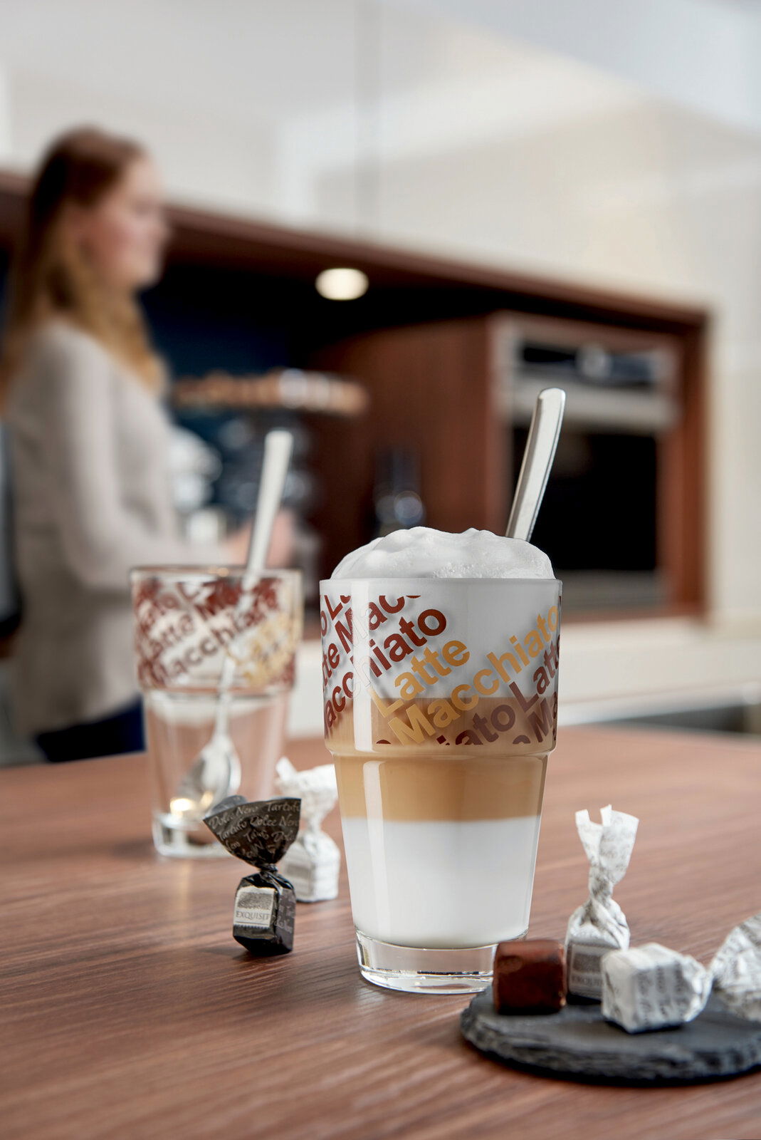 Nahaufnahme eines Cafe Latte Sets mit zwei Gläsern, die mit 'Latte Macchiato' bedruckt sind, und einem Löffel. Im Hintergrund ist eine unscharfe Person in einer modernen Küche zu sehen.