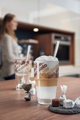 Nahaufnahme eines Cafe Latte Sets mit zwei Gläsern, die mit 'Latte Macchiato' bedruckt sind, und einem Löffel. Im Hintergrund ist eine unscharfe Person in einer modernen Küche zu sehen.