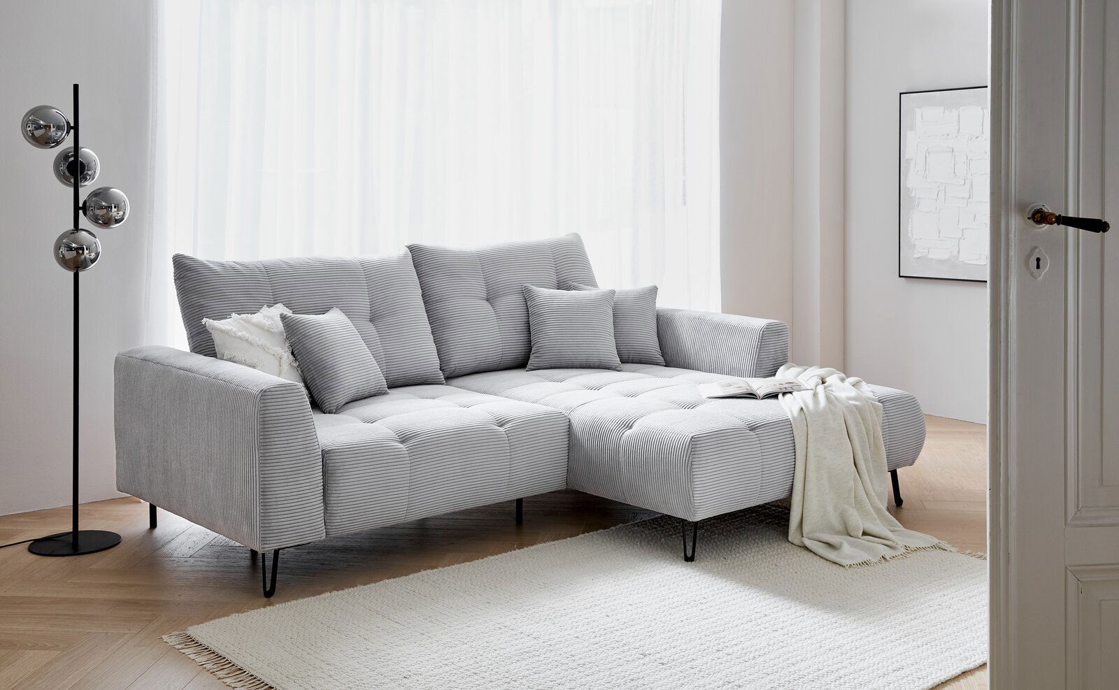 Graues Ecksofa SWING mit Ottomane, dekoriert mit Kissen und Decke, in einem hellen Wohnzimmer, seitliche Perspektive.