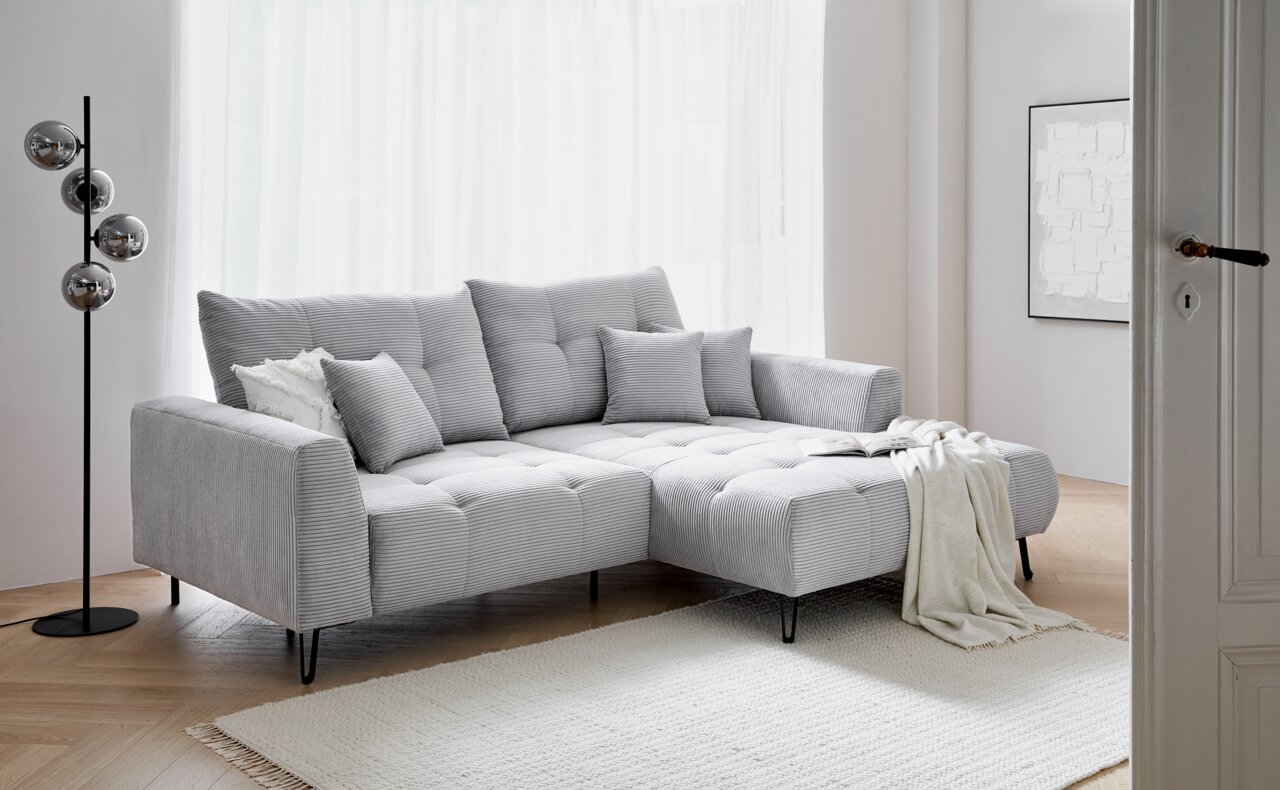 Graues Ecksofa SWING mit Ottomane, dekoriert mit Kissen und Decke, in einem hellen Wohnzimmer, seitliche Perspektive.