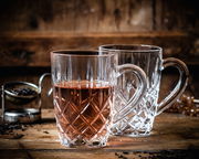 Zwei elegante Teebecher aus Kristallglas mit Henkel, einer gefüllt mit Tee, auf einem rustikalen Holztisch, seitliche Perspektive.