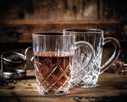 Zwei elegante Teebecher aus Kristallglas mit Henkel, einer gefüllt mit Tee, auf einem rustikalen Holztisch, seitliche Perspektive.