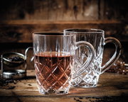 Zwei elegante Teebecher aus Kristallglas mit Henkel, einer gefüllt mit Tee, auf einem rustikalen Holztisch, seitliche Perspektive.