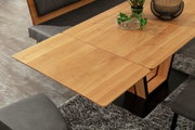 Houten eettafel met uitschuifbaar blad, gefotografeerd van bovenaf, omringd door grijze stoelen en een tapijt.