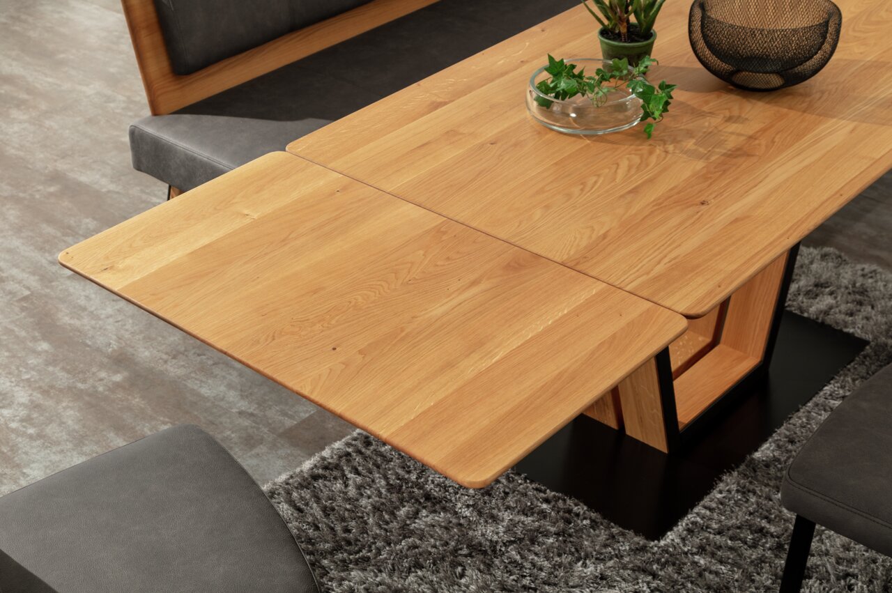 Houten eettafel met uitschuifbaar blad, gefotografeerd van bovenaf, omringd door grijze stoelen en een tapijt.